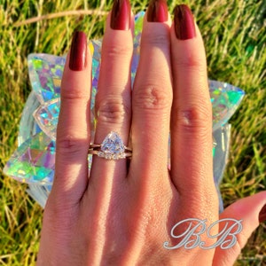 May include: A close-up of a hand wearing a gold ring with a large, triangular, clear gemstone. The ring is set with smaller, clear gemstones around the main stone. The hand is wearing a second, thinner gold band.