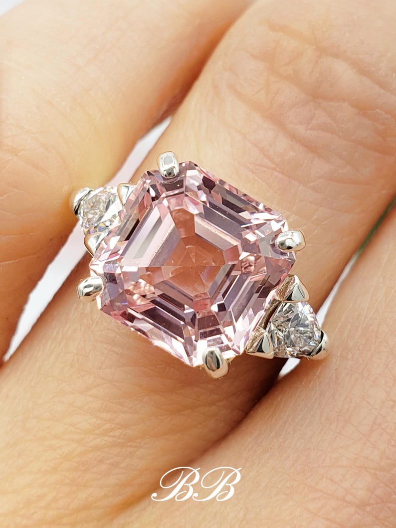 Puede incluir: Primer plano de un anillo con una gran gema rosa claro de talla cuadrada con gemas blancas triangulares m&aacute;s peque&ntilde;as a cada lado. El anillo est&aacute; engastado en una banda plateada. Las iniciales "BB" est&aacute;n en la parte inferior.