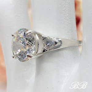 May include: A silver ring featuring a large, round, clear gemstone as the primary focal point. Two smaller, clear gemstones flank the main stone. The ring is set on a white surface, with the initials "BB" visible.