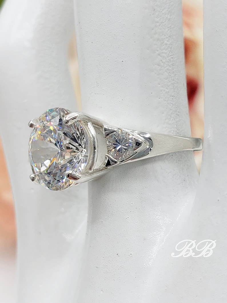 May include: A silver ring featuring a large, round, clear gemstone as the primary focal point. Two smaller, clear gemstones flank the main stone. The ring is set on a white surface, with the initials "BB" visible.
