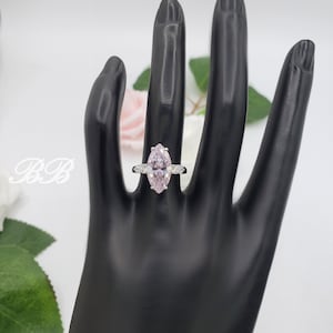 K&ouml;nnte beinhalten: Ein silberner Ring mit einem gro&szlig;en, birnenf&ouml;rmigen, hellrosa Edelstein, der von kleineren, klaren Edelsteinen umgeben ist.