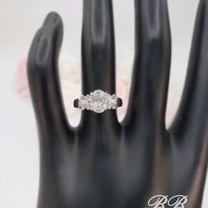 May include: A silver ring with an oval-shaped center stone and smaller stones on the band. The ring is on a black hand model.