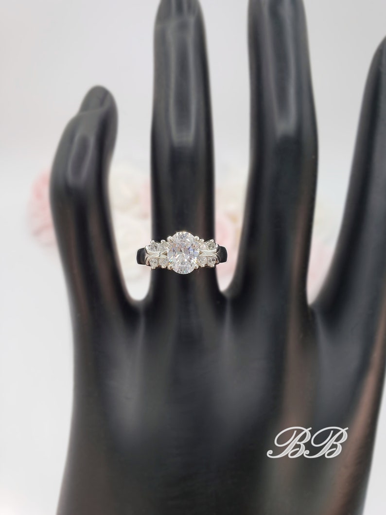 May include: A silver ring with an oval-shaped center stone and smaller stones on the band. The ring is on a black hand model.