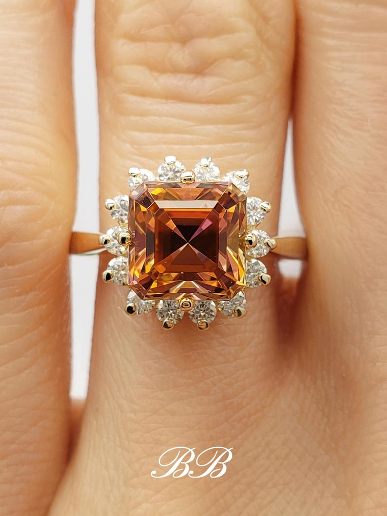 Pode incluir: Um anel de ouro com uma grande pedra preciosa quadrada de cor laranja e marrom, cercada por um halo de pedras preciosas menores e transparentes.