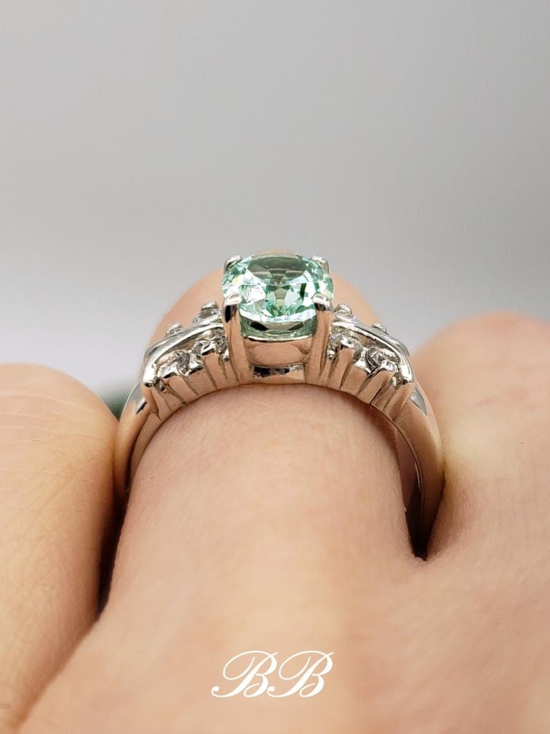 Puede incluir: Un anillo de plata con una piedra preciosa verde claro y piedras preciosas m&aacute;s peque&ntilde;as transparentes engastadas en una banda.
