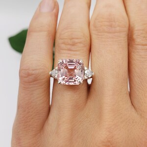 Puede incluir: Un anillo de piedra preciosa rosa con una gran piedra central rectangular y dos piedras laterales triangulares m&aacute;s peque&ntilde;as. El anillo est&aacute; engastado en una banda plateada. Las iniciales "BB" est&aacute;n en la parte inferior.