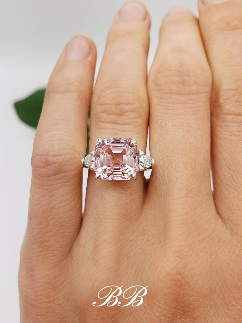 Puede incluir: Un anillo de piedra preciosa rosa con una gran piedra central rectangular y dos piedras laterales triangulares m&aacute;s peque&ntilde;as. El anillo est&aacute; engastado en una banda plateada. Las iniciales "BB" est&aacute;n en la parte inferior.
