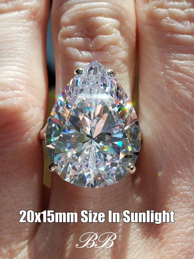 May include: A pear-shaped clear crystal ring with a silver band. The ring is 20x15mm in size and is shown in sunlight.