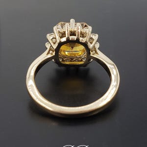 Pode incluir: Um anel de ouro com uma grande gema amarela de forma quadrada cercada por gemas brancas menores. O anel tem uma banda simples.