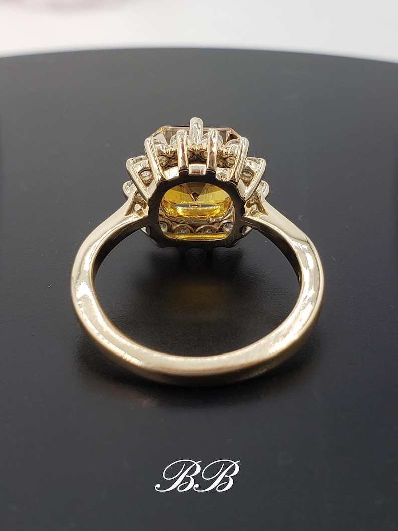 Pode incluir: Um anel de ouro com uma grande gema amarela de forma quadrada cercada por gemas brancas menores. O anel tem uma banda simples.