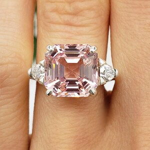 Puede incluir: Primer plano de un anillo con una gran piedra preciosa rosa claro con talla esmeralda como pieza central. El anillo tiene una banda plateada y est&aacute; flanqueado por dos piedras blancas m&aacute;s peque&ntilde;as en forma de pera. Las iniciales "BB" est&aacute;n en la parte inferior.
