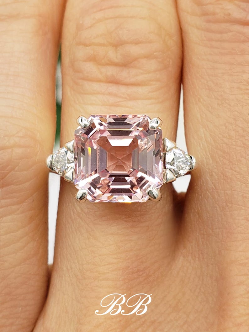 Puede incluir: Primer plano de un anillo con una gran piedra preciosa rosa claro con talla esmeralda como pieza central. El anillo tiene una banda plateada y est&aacute; flanqueado por dos piedras blancas m&aacute;s peque&ntilde;as en forma de pera. Las iniciales "BB" est&aacute;n en la parte inferior.