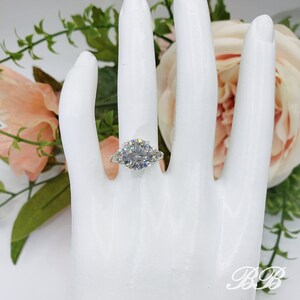 May include: A silver ring with a large, round, clear gemstone as the centerpiece, flanked by two smaller gemstones. The ring is displayed on a white hand, with a floral arrangement in the background.