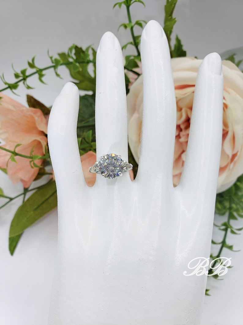 May include: A silver ring with a large, round, clear gemstone as the centerpiece, flanked by two smaller gemstones. The ring is displayed on a white hand, with a floral arrangement in the background.