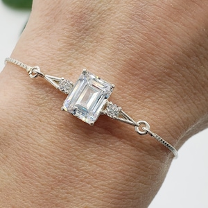 May include: A silver bracelet with a large, clear, emerald-cut cubic zirconia stone and smaller accent stones. The bracelet is on a delicate chain.
