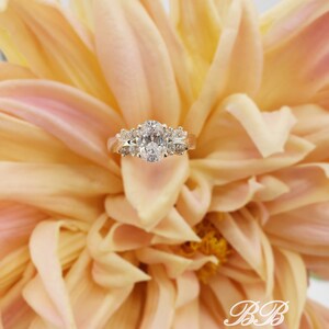 May include: A gold engagement ring with an oval-shaped center diamond and smaller diamonds on the band. The ring is set against a backdrop of soft, peach-colored flower petals.
