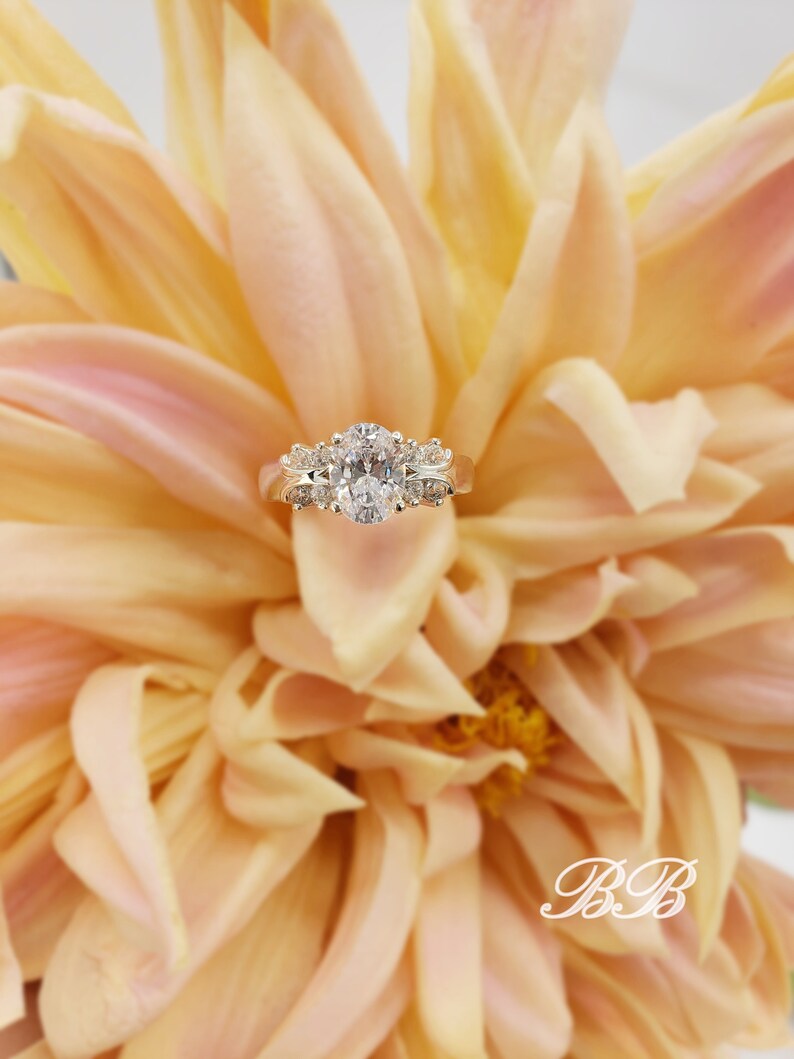 May include: A gold engagement ring with an oval-shaped center diamond and smaller diamonds on the band. The ring is set against a backdrop of soft, peach-colored flower petals.