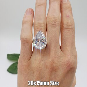 May include: A large pear-shaped clear cubic zirconia ring set in a gold band. The ring is on a person's finger. The text "20x15mm Size" is written below the ring. The letters "B.B" are written below the text.