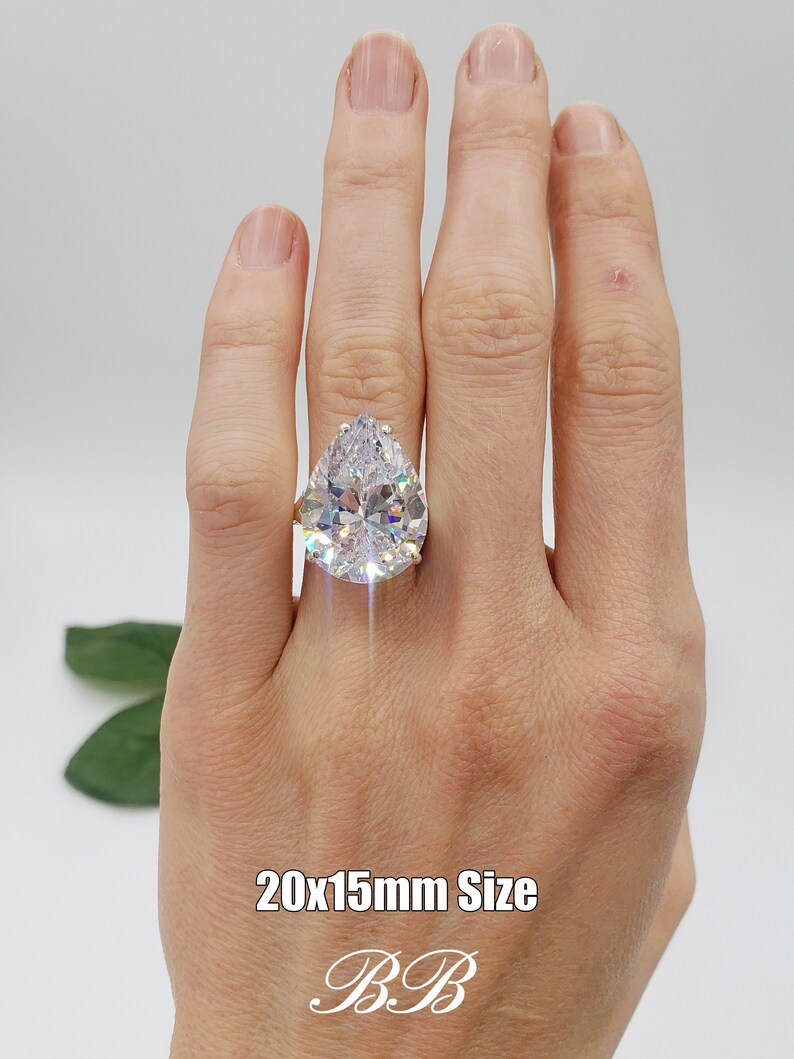 May include: A large pear-shaped clear cubic zirconia ring set in a gold band. The ring is on a person's finger. The text "20x15mm Size" is written below the ring. The letters "B.B" are written below the text.