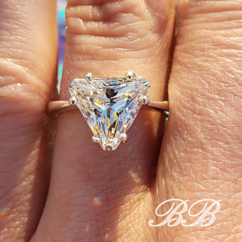 May include: A rose gold ring featuring a large, clear, triangular-cut gemstone. The gemstone is held in place by four prongs. The ring is shown on a finger, with the initials "BB" in the lower right corner.