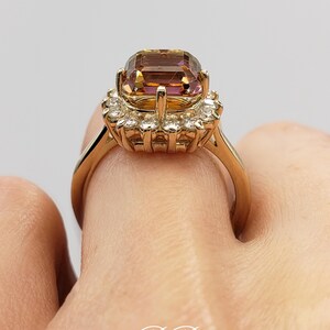 Pode incluir: Um anel de ouro com uma grande gema quadrada, de cor roxa e marrom, cercada por um halo de gemas menores e transparentes.