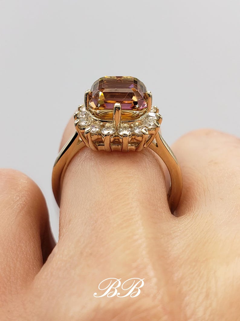 Pode incluir: Um anel de ouro com uma grande gema quadrada, de cor roxa e marrom, cercada por um halo de gemas menores e transparentes.