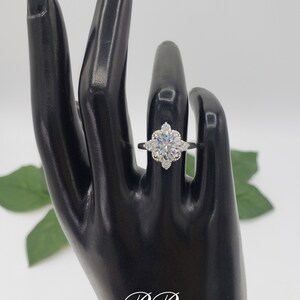 Puede incluir: Un anillo de plata con una piedra central redonda grande y clara, rodeada de piedras m&aacute;s peque&ntilde;as y claras. El anillo est&aacute; en un maniqu&iacute; de mano negro.
