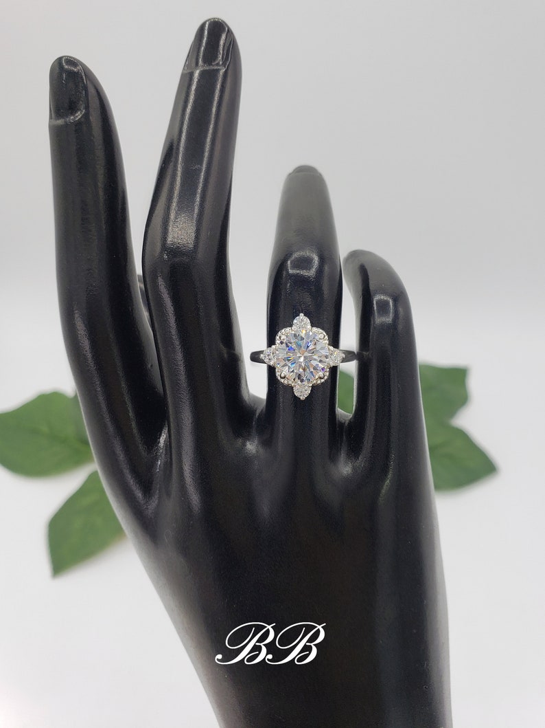 Puede incluir: Un anillo de plata con una piedra central redonda grande y clara, rodeada de piedras m&aacute;s peque&ntilde;as y claras. El anillo est&aacute; en un maniqu&iacute; de mano negro.