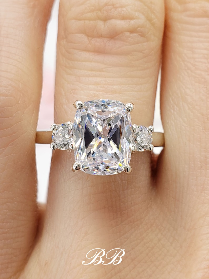 May include: A silver ring with a large, rectangular, clear, cushion-cut center stone and two smaller round clear stones on either side of the center stone.