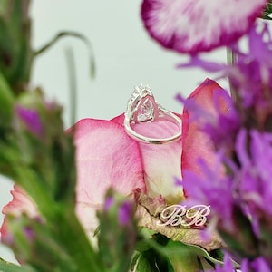 May include: A silver ring with a pear-shaped clear stone set in a halo of smaller stones. The ring is resting on a pink flower petal.