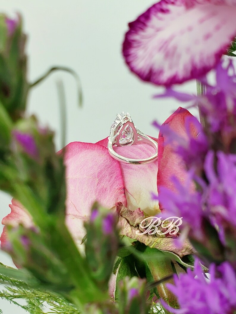 May include: A silver ring with a pear-shaped clear stone set in a halo of smaller stones. The ring is resting on a pink flower petal.