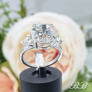 May include: A silver ring with a large, round, clear gemstone as the centerpiece, flanked by two smaller, triangular stones. The ring is displayed on a black stand against a soft, blurred floral background. The ring's band is smooth and polished.