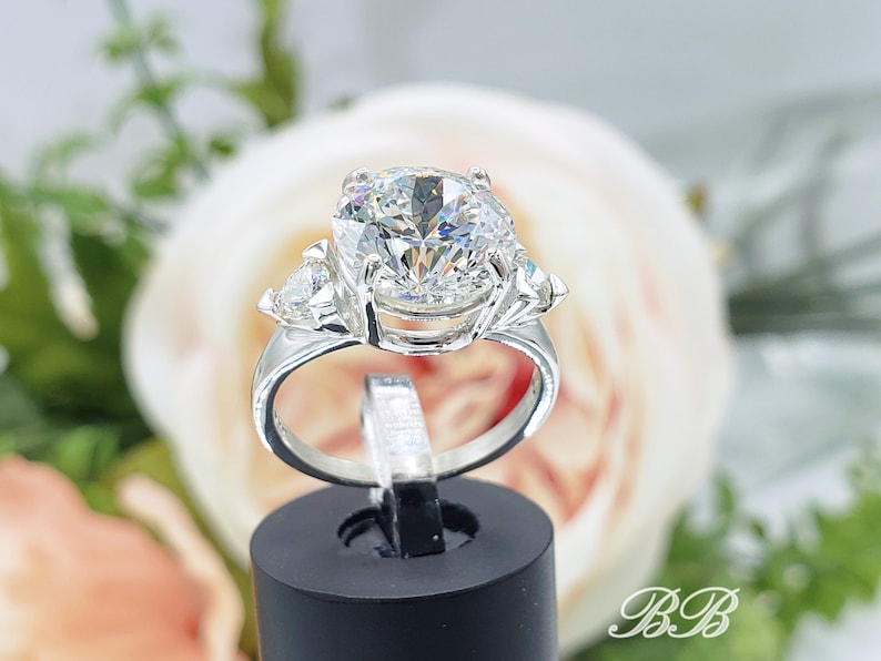 May include: A silver ring with a large, round, clear gemstone as the centerpiece, flanked by two smaller, triangular stones. The ring is displayed on a black stand against a soft, blurred floral background. The ring's band is smooth and polished.