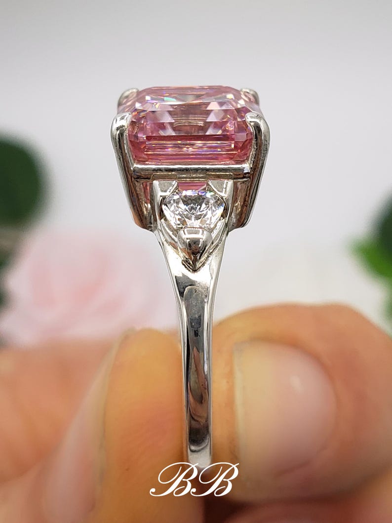 Puede incluir: Un anillo plateado con una gran gema rosa rectangular como elemento principal. Una piedra transparente m&aacute;s peque&ntilde;a en forma de coraz&oacute;n est&aacute; engastada debajo de la piedra principal. La banda del anillo es delgada y pulida, con las iniciales "BB" en escritura.