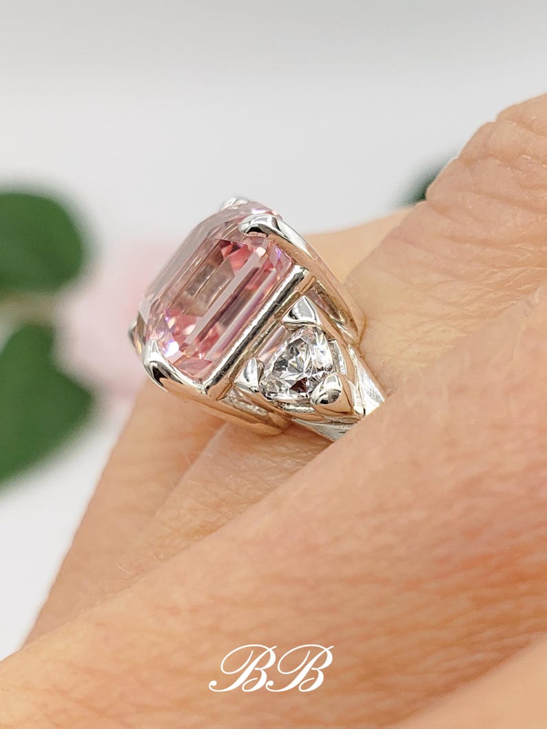 Puede incluir: Un anillo de plata con una gran gema rosa rectangular como pieza central. Dos gemas claras triangulares m&aacute;s peque&ntilde;as flanquean la piedra principal. El anillo tiene un dise&ntilde;o detallado. Las iniciales "BB" est&aacute;n en la parte inferior.