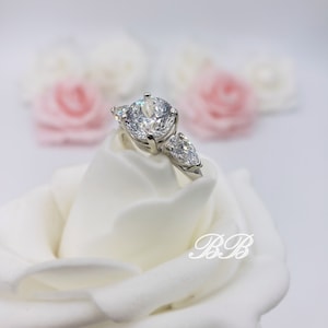 May include: A silver ring with a large round center stone and two smaller pear-shaped stones on either side. The ring is displayed on a white rose.