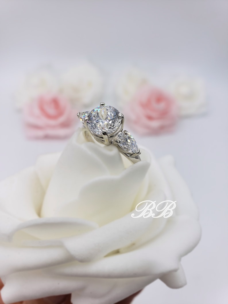 May include: A silver ring with a large round center stone and two smaller pear-shaped stones on either side. The ring is displayed on a white rose.