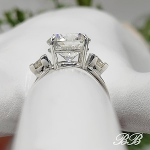 May include: A three-stone engagement ring with a large, clear, square-cut center stone and two smaller, triangular side stones. The ring is set in a silver band, showcasing the brilliance of the diamonds. The design is classic and elegant.