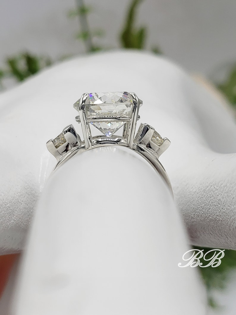 May include: A three-stone engagement ring with a large, clear, square-cut center stone and two smaller, triangular side stones. The ring is set in a silver band, showcasing the brilliance of the diamonds. The design is classic and elegant.