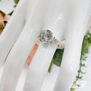 May include: A silver ring with a large, round, clear gemstone as the centerpiece, flanked by two smaller triangular stones. The ring is set on a white surface, with floral accents in the background.