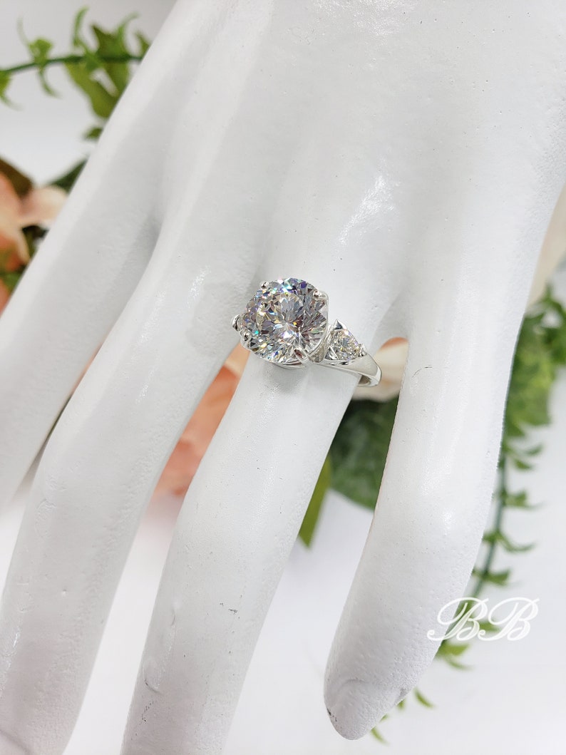 May include: A silver ring with a large, round, clear gemstone as the centerpiece, flanked by two smaller triangular stones. The ring is set on a white surface, with floral accents in the background.