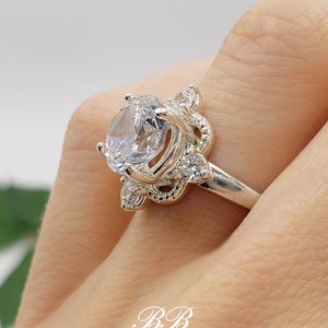 Puede incluir: Un anillo de plata con una piedra grande, transparente y de corte redondo rodeada de piedras m&aacute;s peque&ntilde;as. El anillo tiene un dise&ntilde;o delicado y ornamentado.