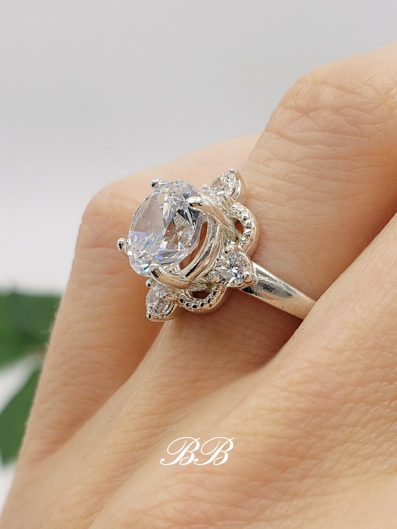 Puede incluir: Un anillo de plata con una piedra grande, transparente y de corte redondo rodeada de piedras m&aacute;s peque&ntilde;as. El anillo tiene un dise&ntilde;o delicado y ornamentado.