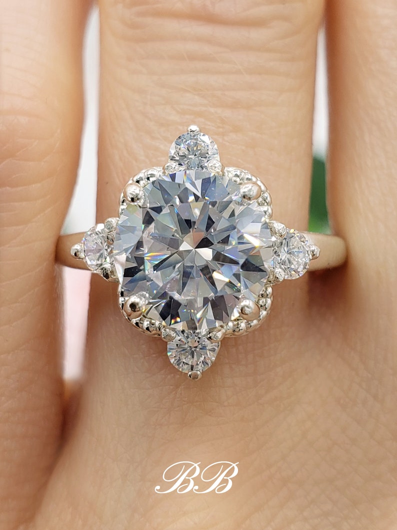 May include: A silver ring with a large, clear, round-cut center stone surrounded by smaller, clear, round-cut stones. The ring has a delicate, scalloped design.