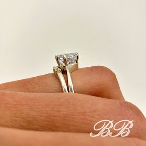 May include: A silver ring set with a large, clear, round-cut cubic zirconia stone. The engagement ring has a delicate band with smaller cubic zirconia stones on the side. The wedding band is a simple, plain band.