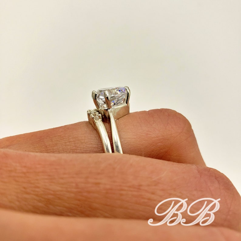 May include: A silver ring set with a large, clear, round-cut cubic zirconia stone. The engagement ring has a delicate band with smaller cubic zirconia stones on the side. The wedding band is a simple, plain band.