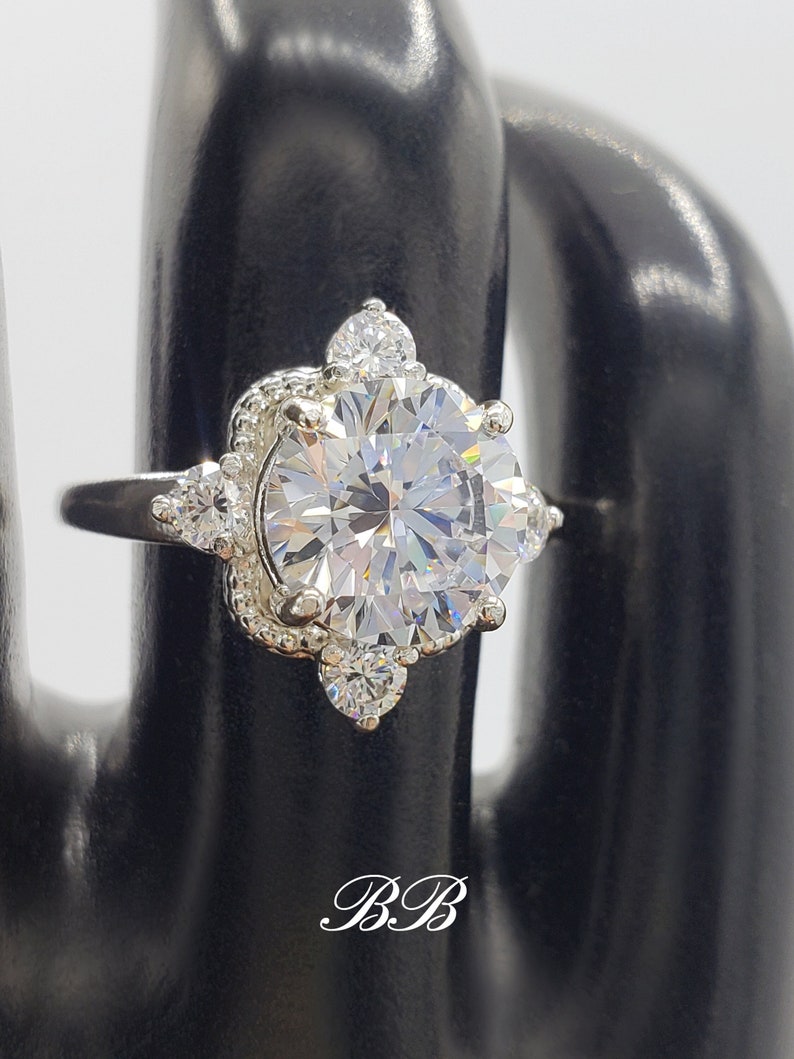 May include: A silver ring with a large round clear stone surrounded by smaller clear stones. The ring is set in a decorative silver setting. The ring is on a black ring display stand. The stand has the letters 'B&B' written on it.