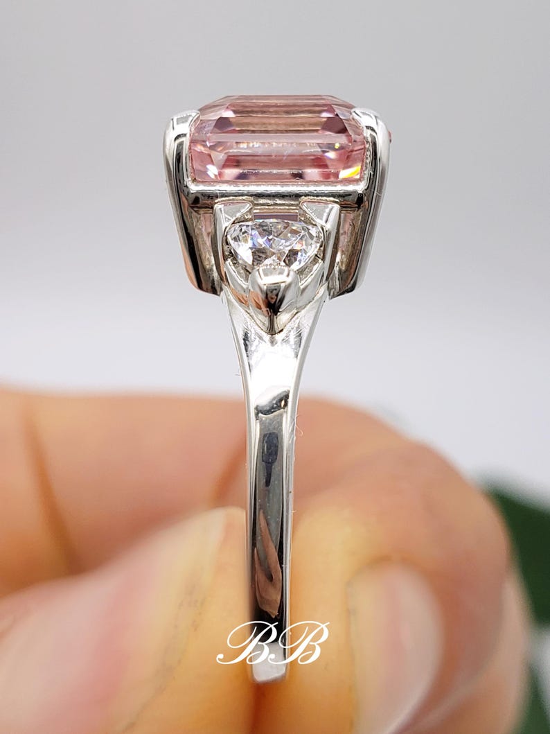Puede incluir: Un anillo de plata con una gran gema rosa rectangular. Una piedra m&aacute;s peque&ntilde;a, transparente, en forma de coraz&oacute;n est&aacute; engastada debajo de la piedra principal. El anillo tiene una superficie pulida y reflectante. Las iniciales "BB" est&aacute;n en la base del anillo.