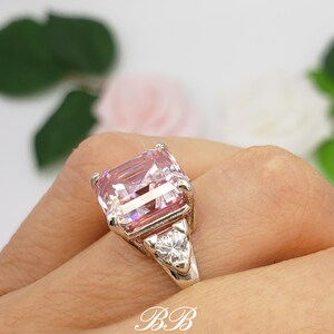 Puede incluir: Un anillo de plata con una gran gema rosa rectangular y dos piedras claras triangulares m&aacute;s peque&ntilde;as. El anillo est&aacute; en una mano, con las iniciales "BB" visibles en la imagen.