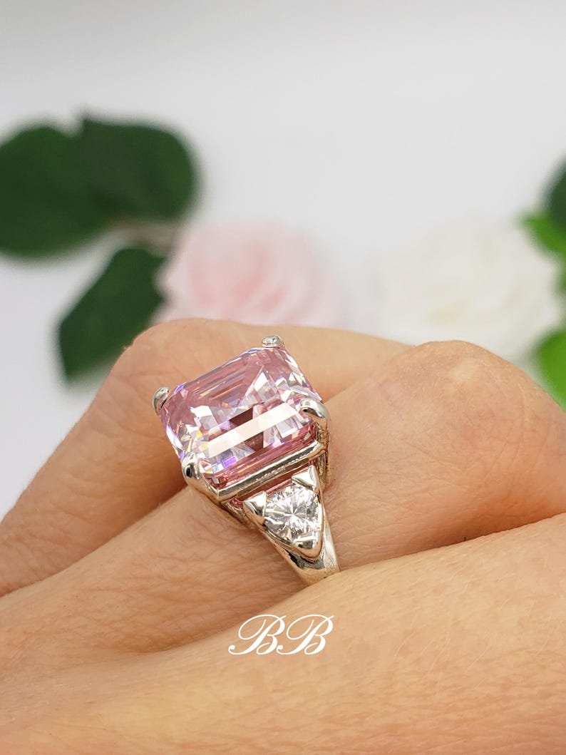Puede incluir: Un anillo de plata con una gran gema rosa rectangular y dos piedras claras triangulares m&aacute;s peque&ntilde;as. El anillo est&aacute; en una mano, con las iniciales "BB" visibles en la imagen.